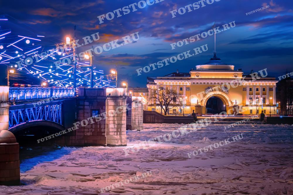 На фотографии запечатлен Дворцовый мост и Адмиралтейство в вечернее время. Мост украшен яркой иллюминацией, отражающейся в замерзшей реке Неве. Ледоход на Неве добавляет особую атмосферу зимнему городскому пейзажу. Вдали виднеется величественное здание Адмиралтейства, освещенное огнями.